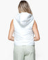 A woman viewed from behind wearing a white sleeveless puffer vest with a hood and light sage green pants, against a plain white studio background.