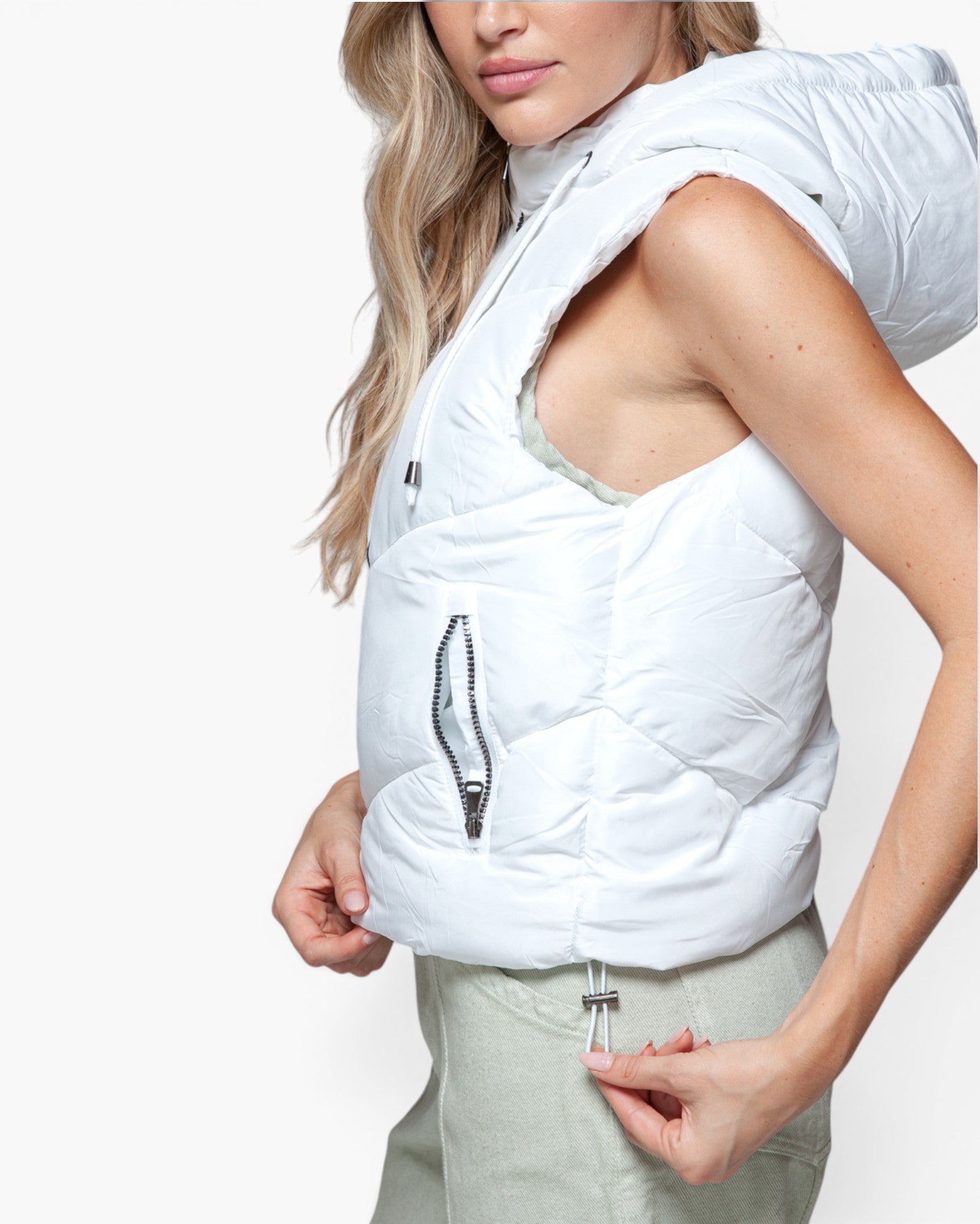 A woman wearing a white sleeveless puffer vest with a hood and silver zipper details, paired with light sage green pants, against a clean white background.