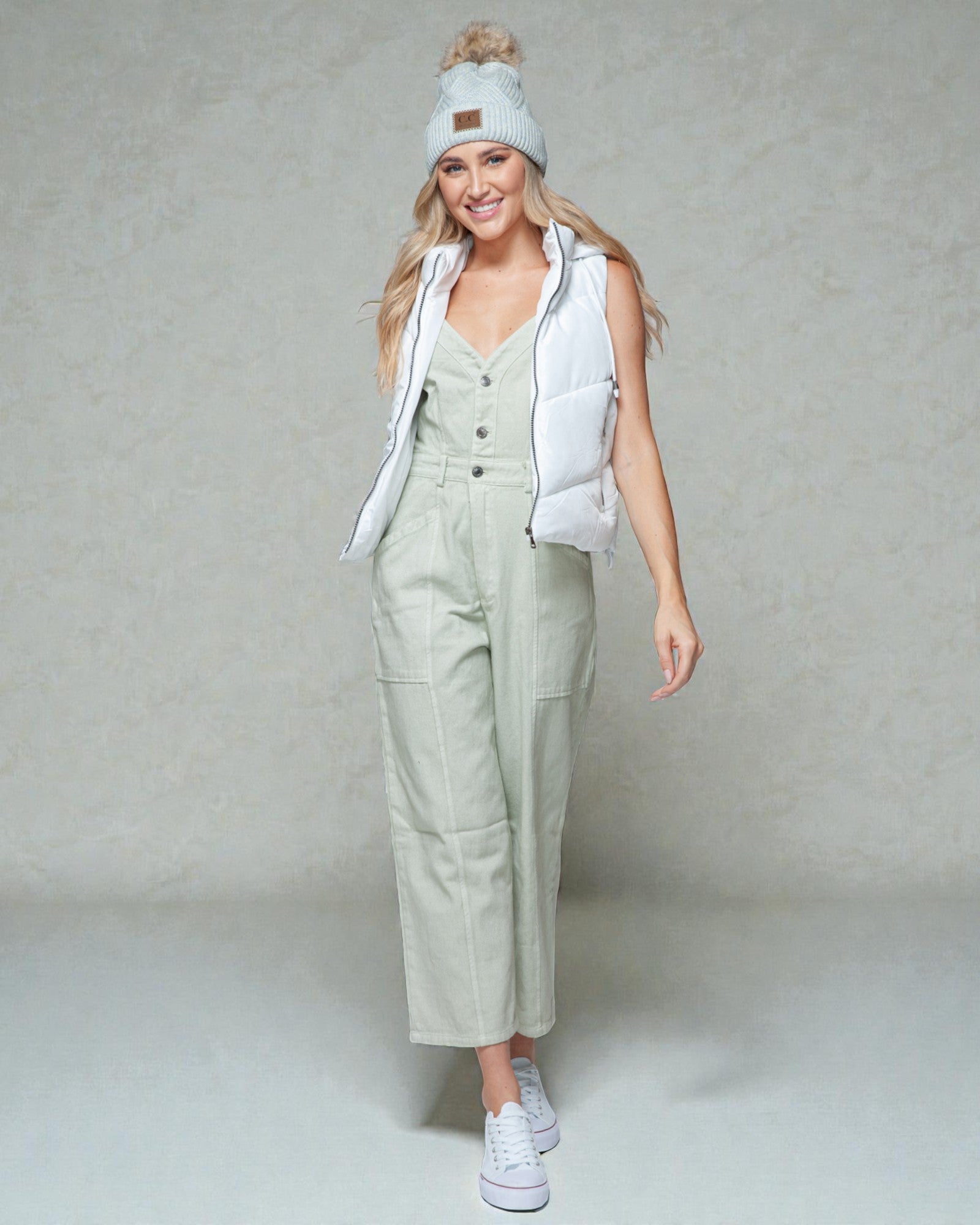 Woman wearing a light green jumpsuit with a white vest and beanie against a plain background