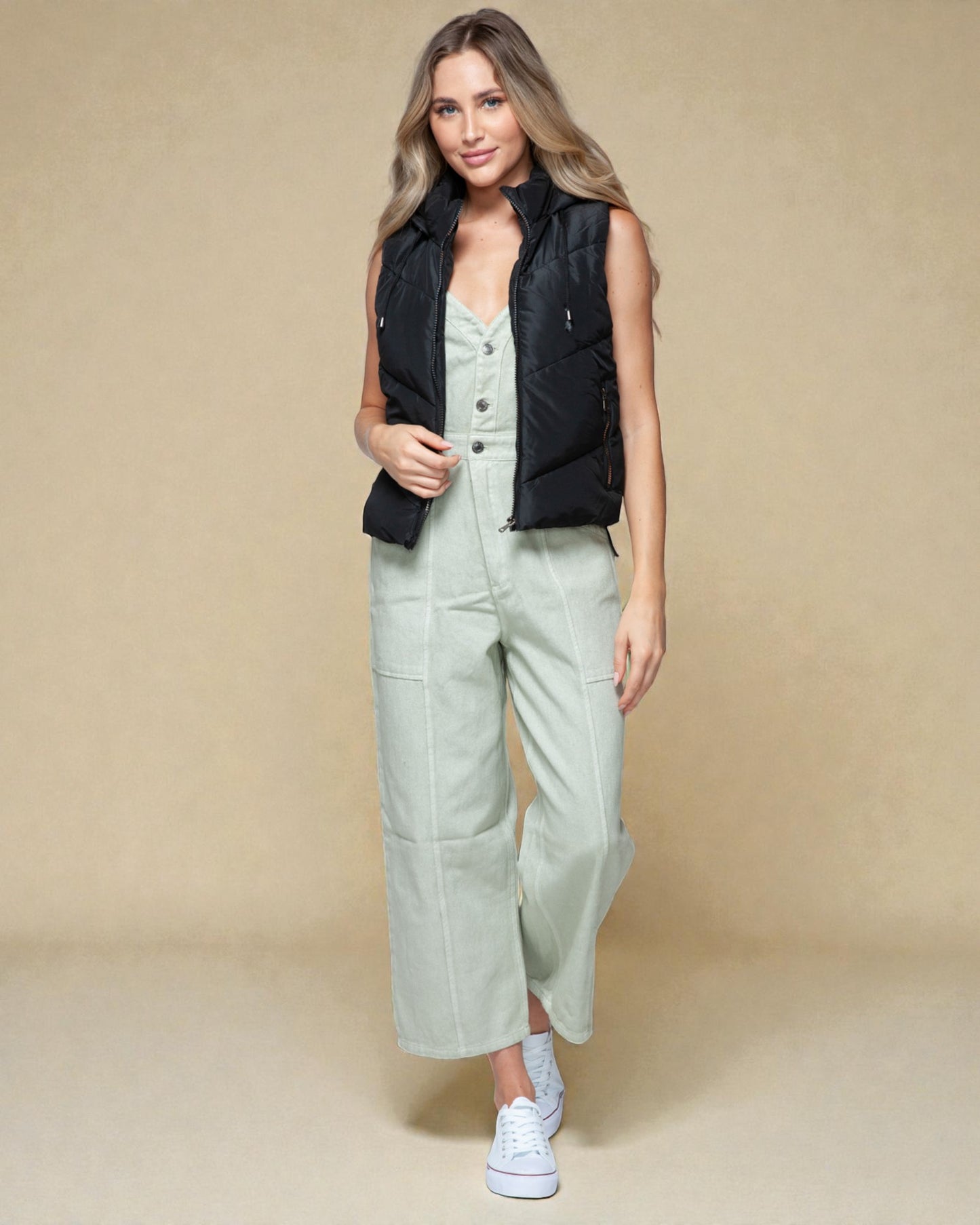 Woman wearing a black vest over a light green dress with white sneakers on a beige background