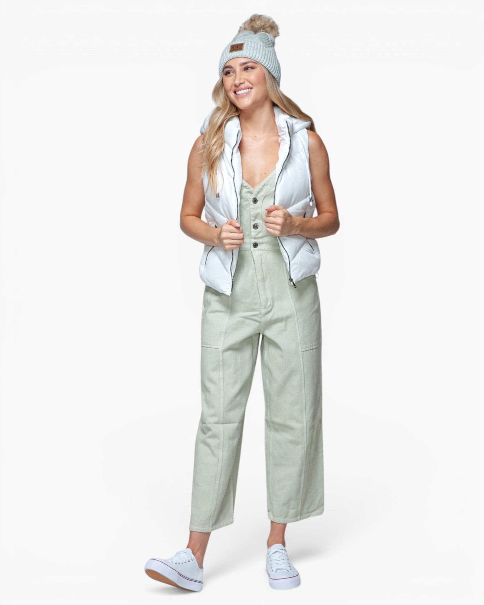 A woman modeling a light sage green utility jumpsuit with a white puffer vest, a light blue knit beanie with a pom-pom, and white sneakers, set against a plain white studio background.