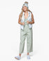 A woman modeling a light sage green utility jumpsuit with a white puffer vest, a light blue knit beanie with a pom-pom, and white sneakers, set against a plain white studio background.