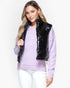 A woman modeling a black shiny puffer vest worn over a light purple knit sweater and white pants, photographed against a clean light gray studio background.
