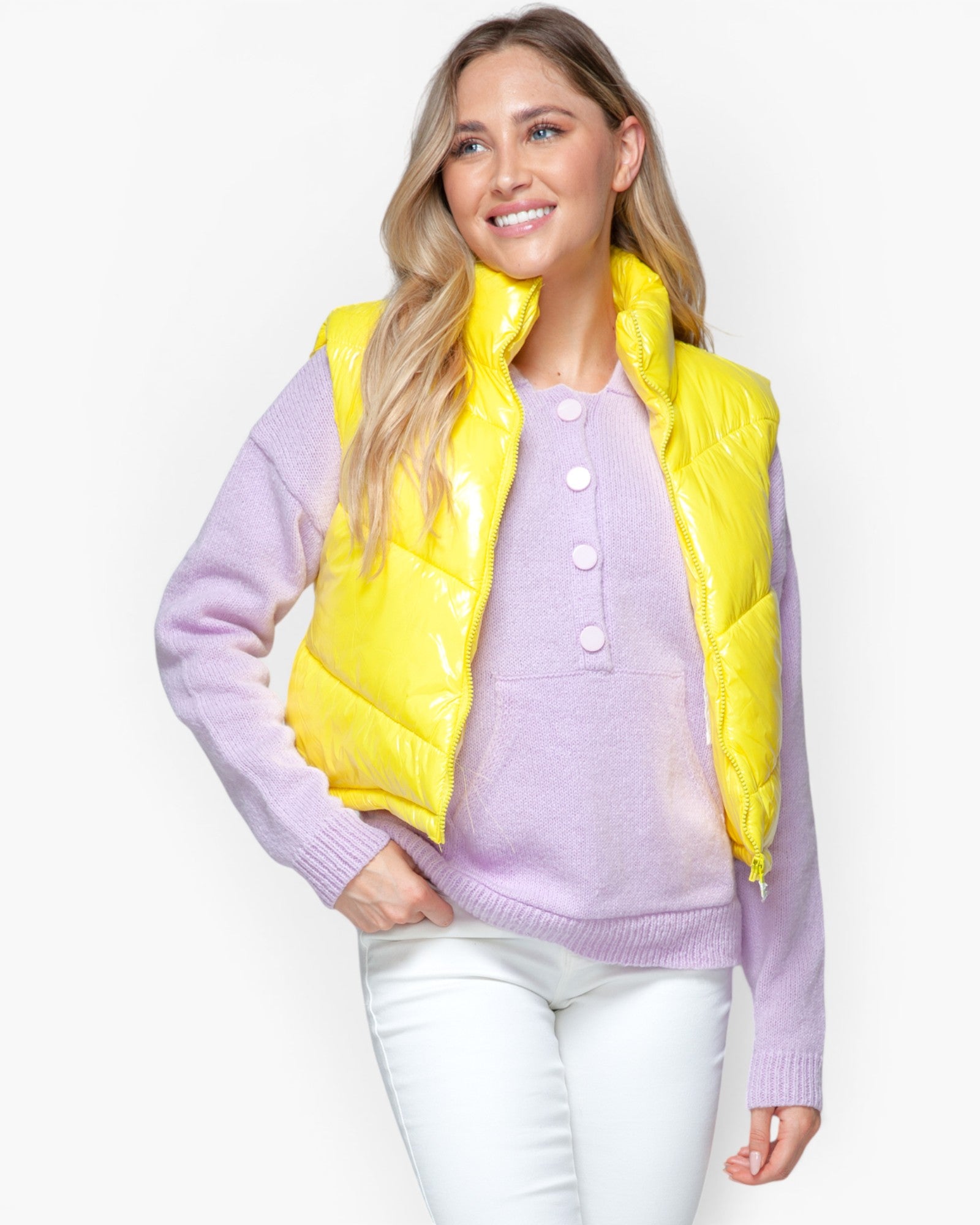 A woman modeling a bright yellow puffer vest over a lavender button-up sweater and white pants, photographed against a clean white studio background.