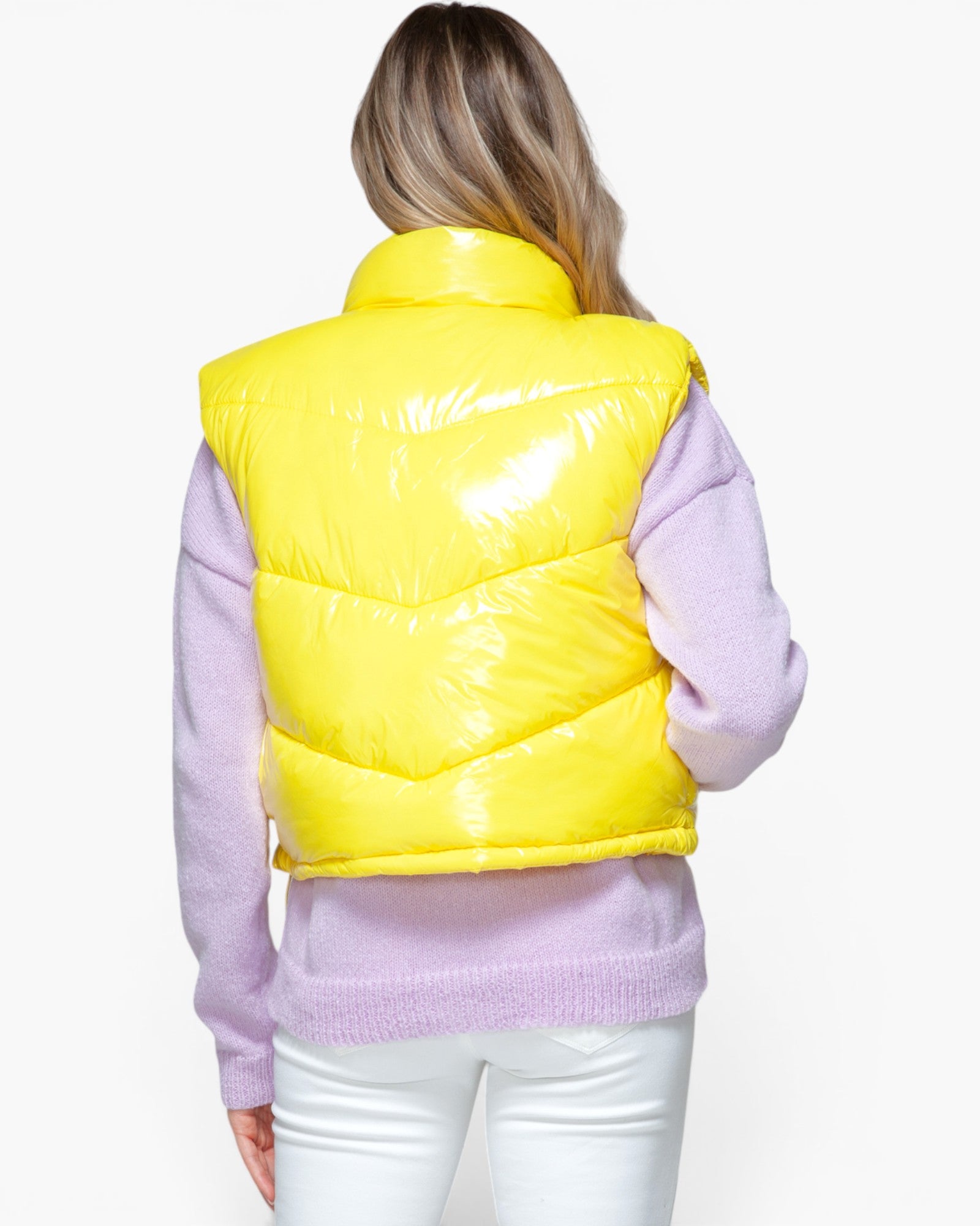 A woman shown from the back wearing a shiny bright yellow puffer vest over a light purple long-sleeve top and white pants, against a plain light gray studio background.