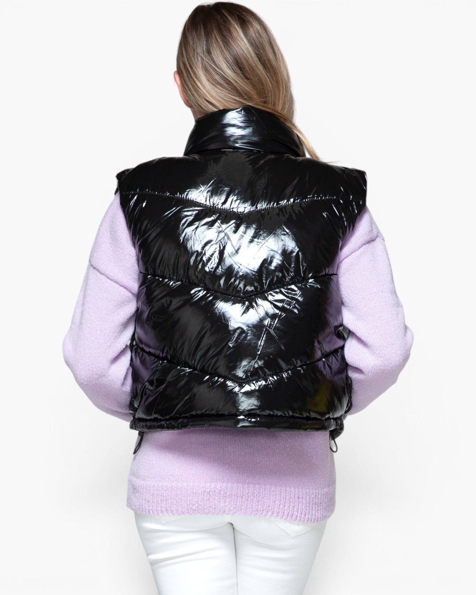 A woman viewed from behind wearing a shiny black puffer vest over a light purple sweater and white pants, against a plain light background.
