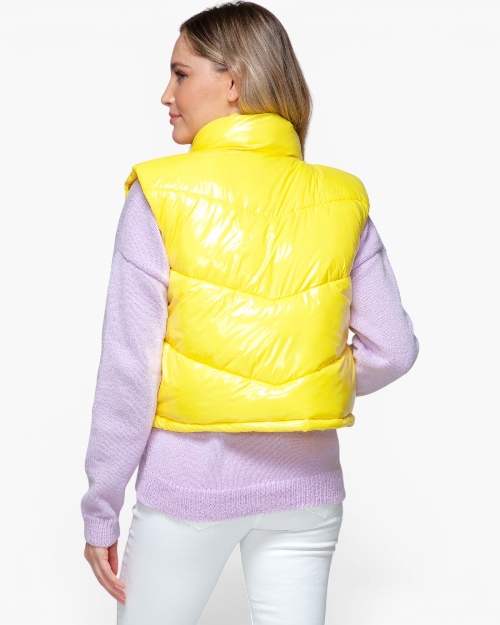 A woman shown from the back wearing a bright yellow shiny puffer vest over a light purple sweater and white pants, against a plain light gray studio background.