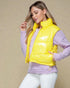 Woman wearing a yellow puffer vest over a purple sweater on a beige background