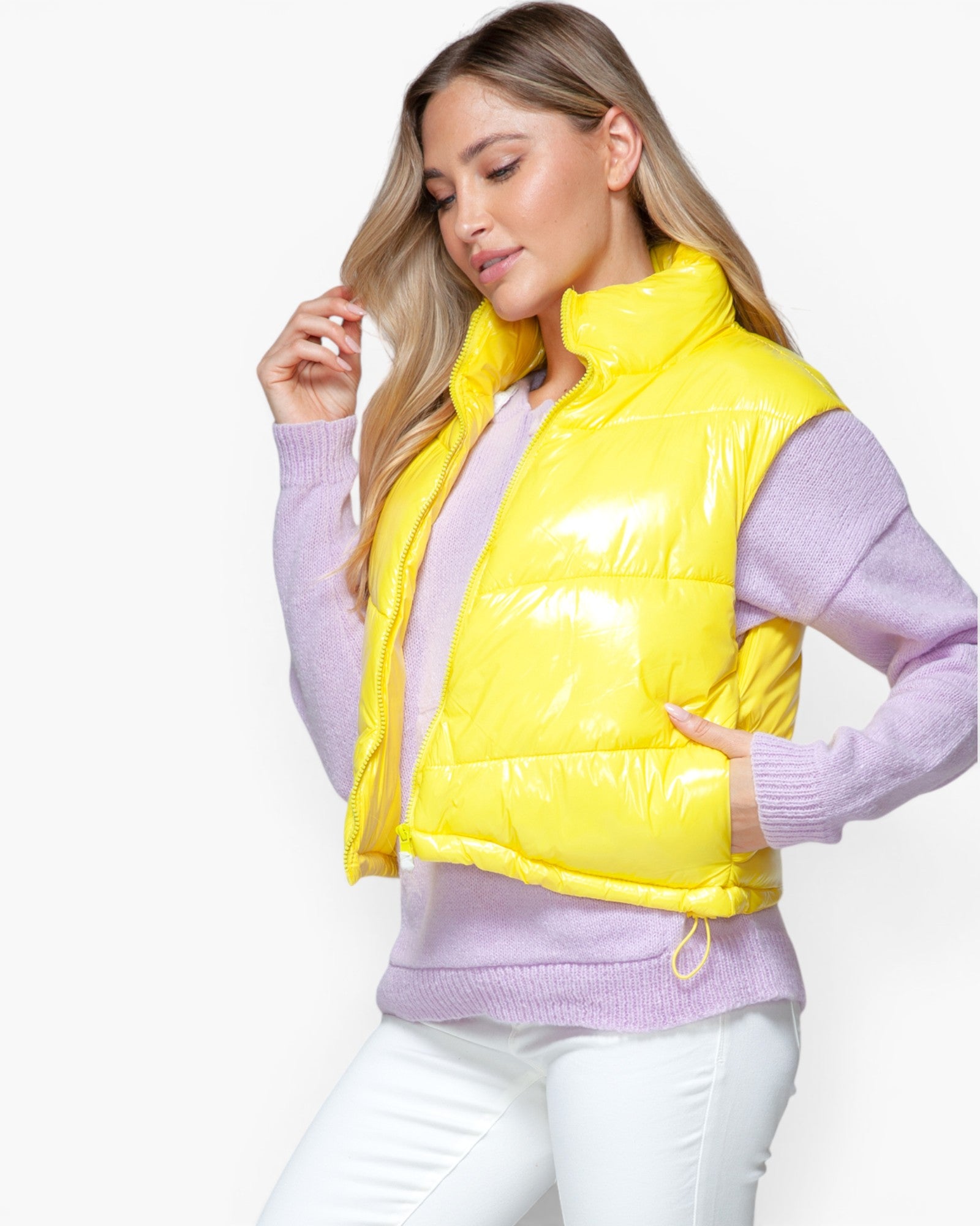 A woman wearing a bright yellow puffer vest over a light purple sweater and white pants, photographed against a clean white studio background.