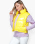 A woman wearing a bright yellow puffer vest over a light purple sweater and white pants, photographed against a clean white studio background.
