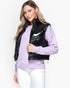 A woman modeling a black shiny puffer vest over a light purple knit sweater and white pants, photographed against a plain light gray studio background.