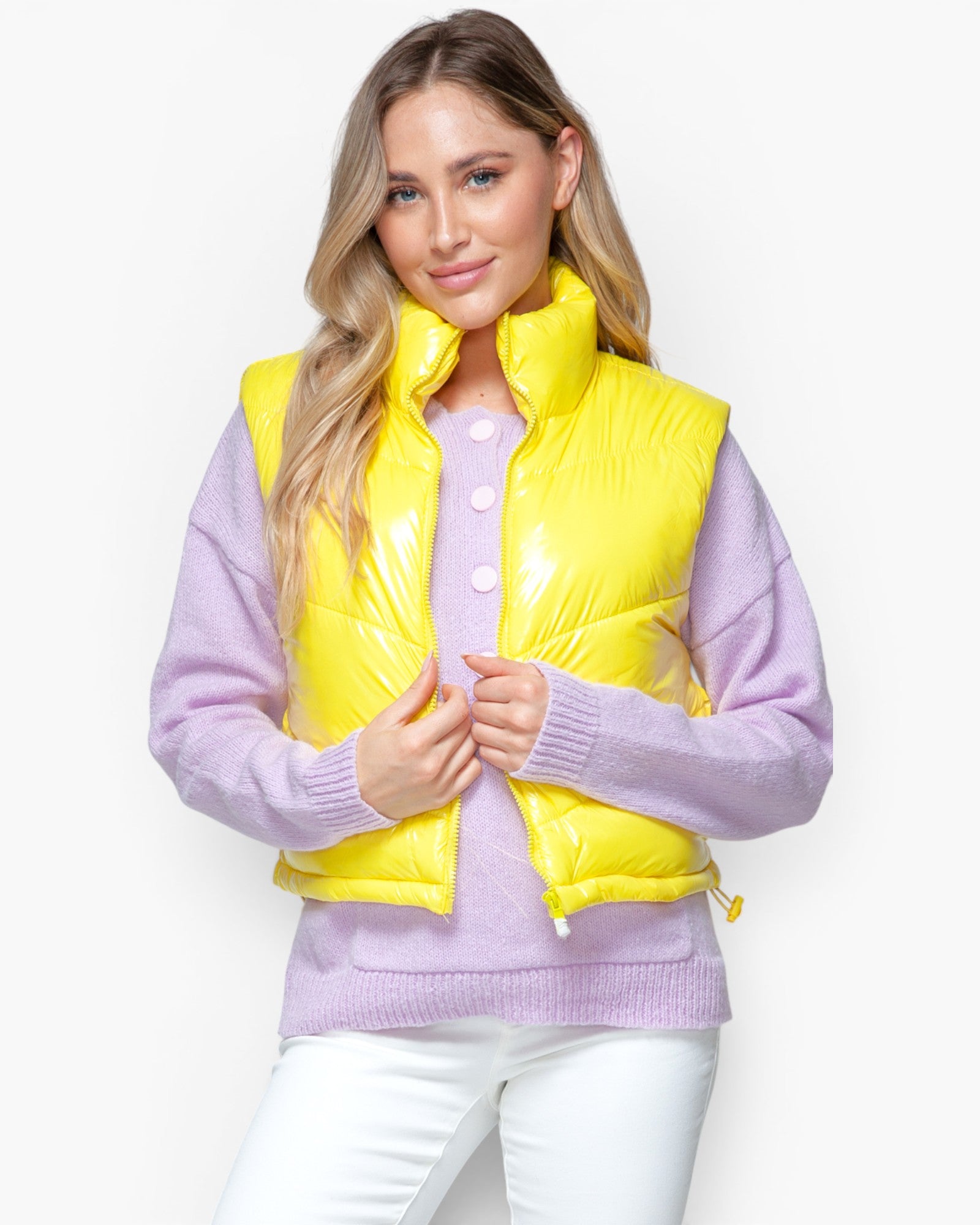 A woman wearing a shiny bright yellow puffer vest over a lavender knit sweater and white pants, photographed against a plain light gray studio background.