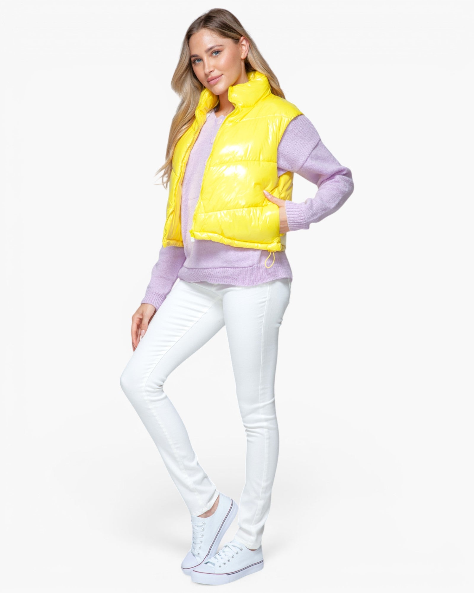 A woman modeling a bright yellow puffer vest over a lavender long-sleeve top, paired with white skinny jeans and white sneakers, against a plain white studio background.