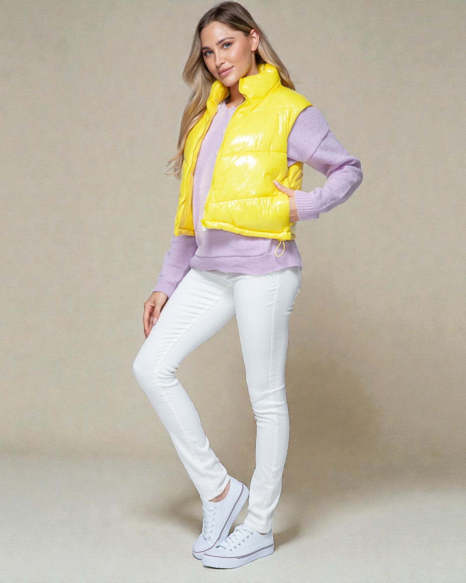 Woman wearing a yellow puffer vest over a purple sweater with white pants on a beige background