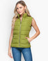 A woman wearing a sleeveless olive green puffer vest with a full front zipper and zippered side pockets, paired with blue jeans, against a light gray studio background.