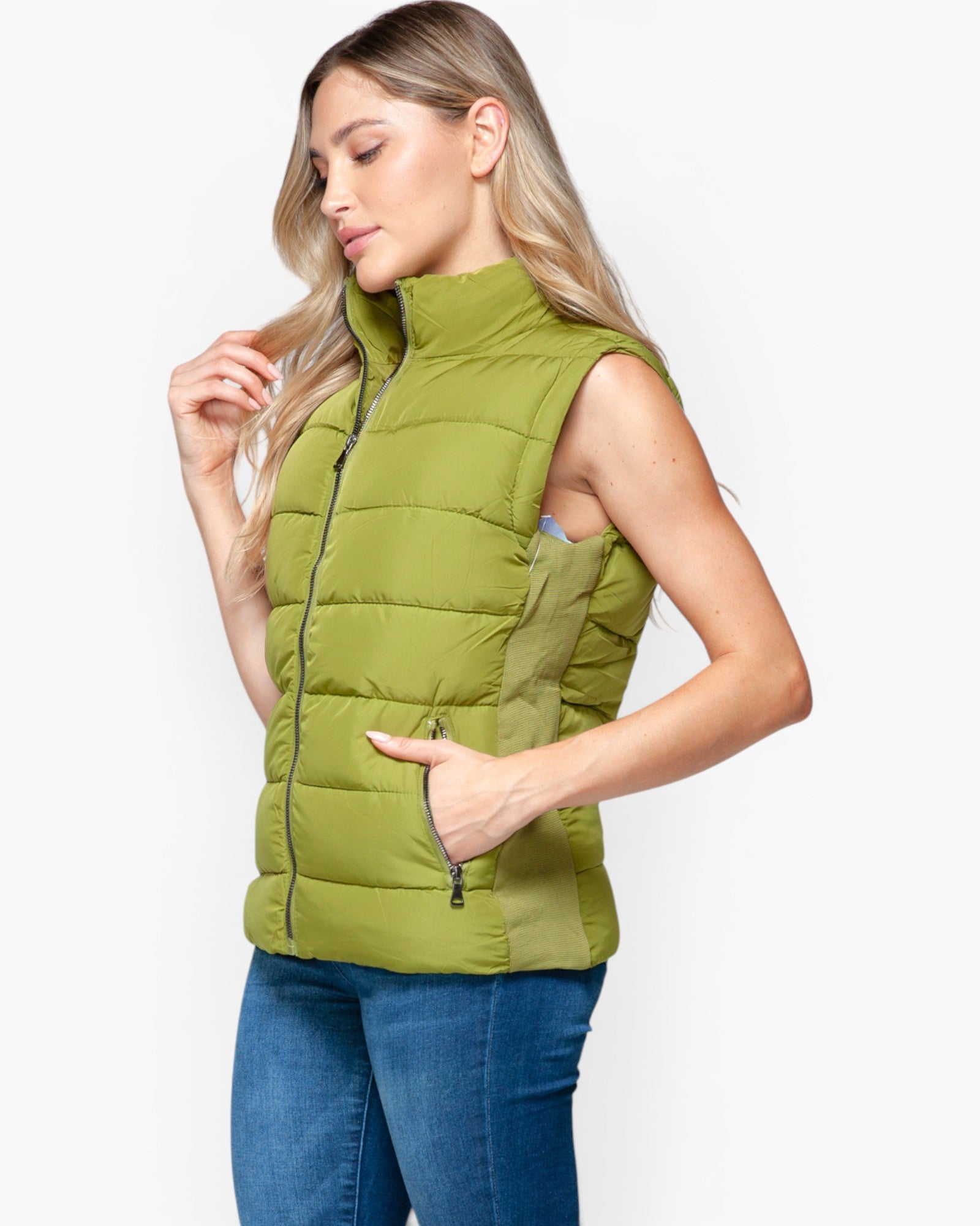 A woman wearing a sleeveless olive green puffer vest with a high collar and front zipper, paired with blue jeans, photographed against a light gray studio background.