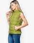 A woman wearing a sleeveless olive green puffer vest with a high collar and front zipper, paired with blue jeans, photographed against a light gray studio background.