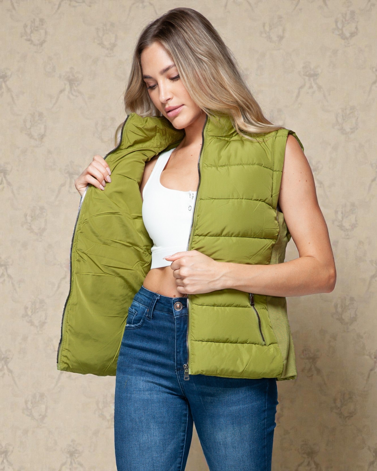Woman wearing a green puffer vest over a white top and blue jeans against a beige wall.