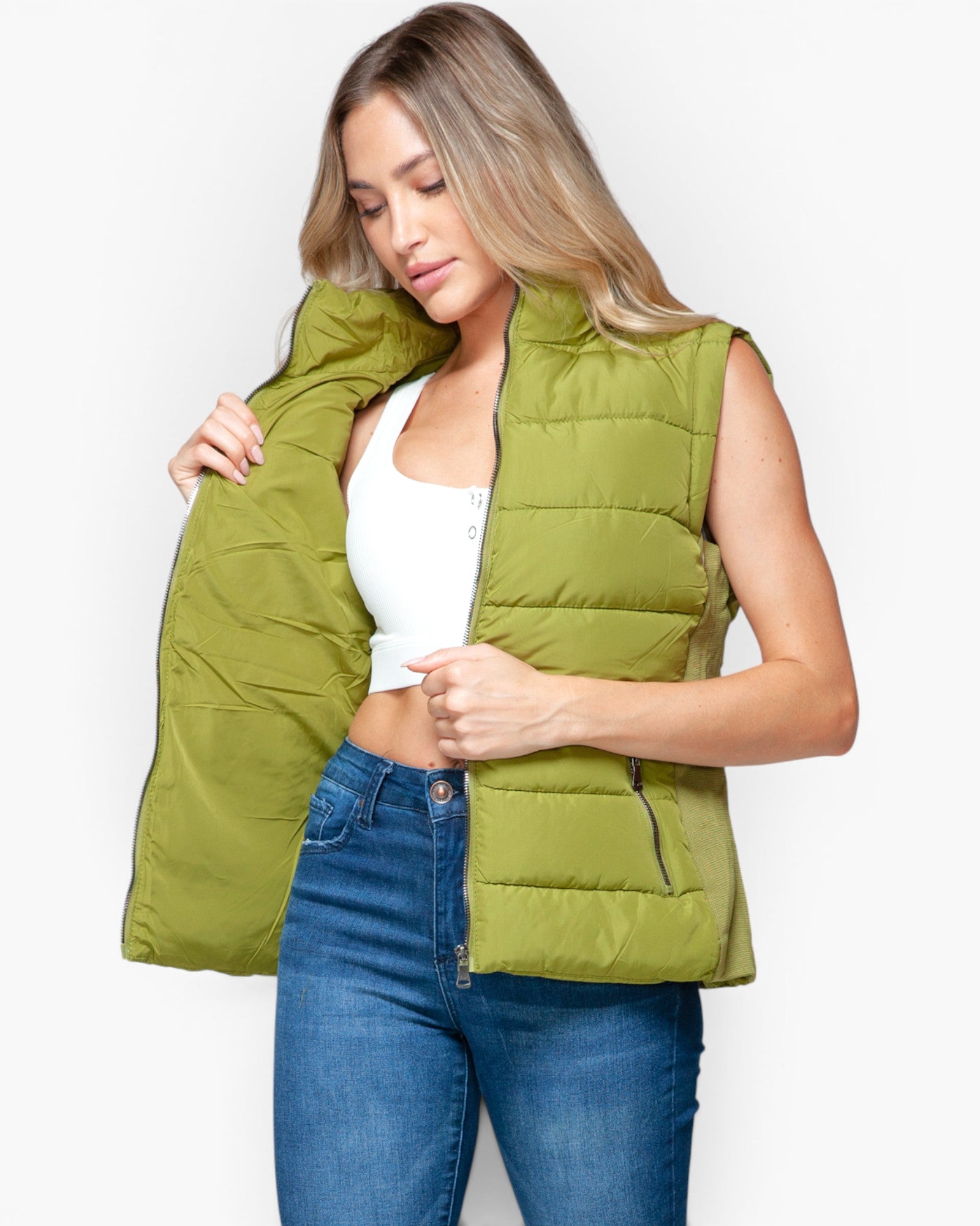 A woman wearing a sleeveless olive green puffer vest over a white crop top and blue jeans, holding the vest open against a plain light gray background.