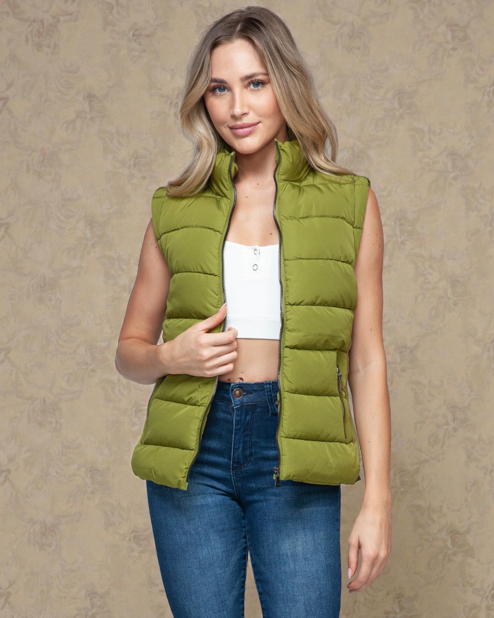 Woman wearing a green puffer vest over a white top and blue jeans on a beige background