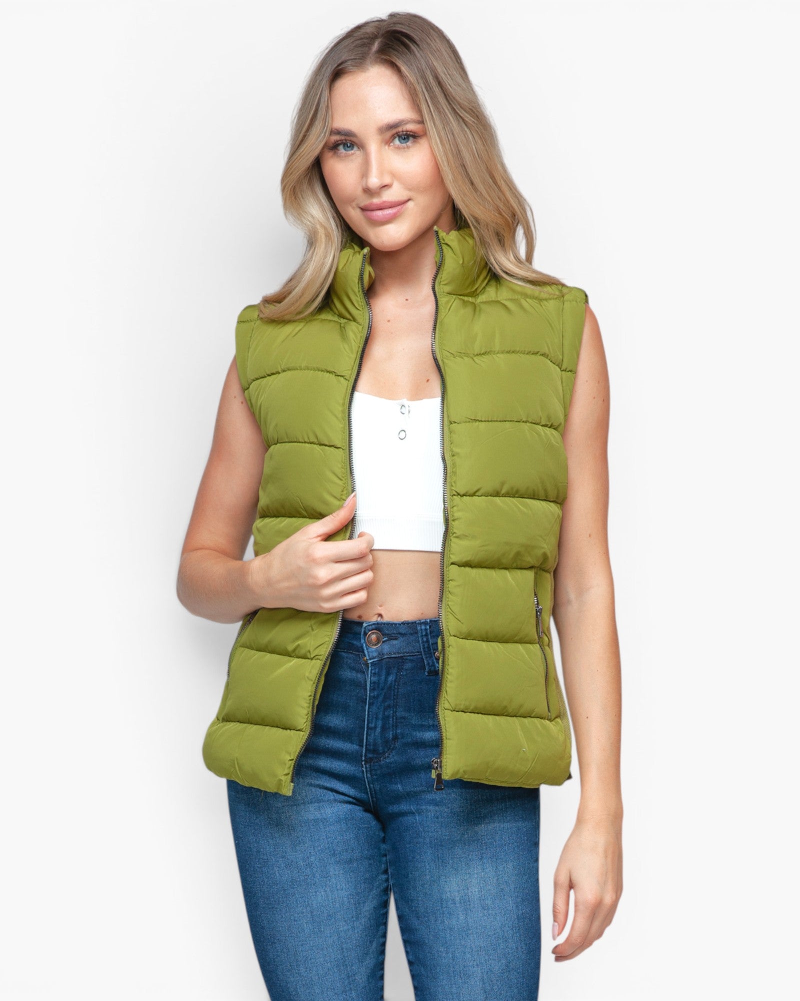 A woman modeling a sleeveless olive green puffer vest over a white crop top and blue jeans, standing against a light gray studio background.