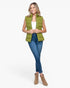 A woman modeling a sleeveless olive green puffer vest over a white top, blue skinny jeans, and tan ankle boots, photographed against a plain light gray studio background.