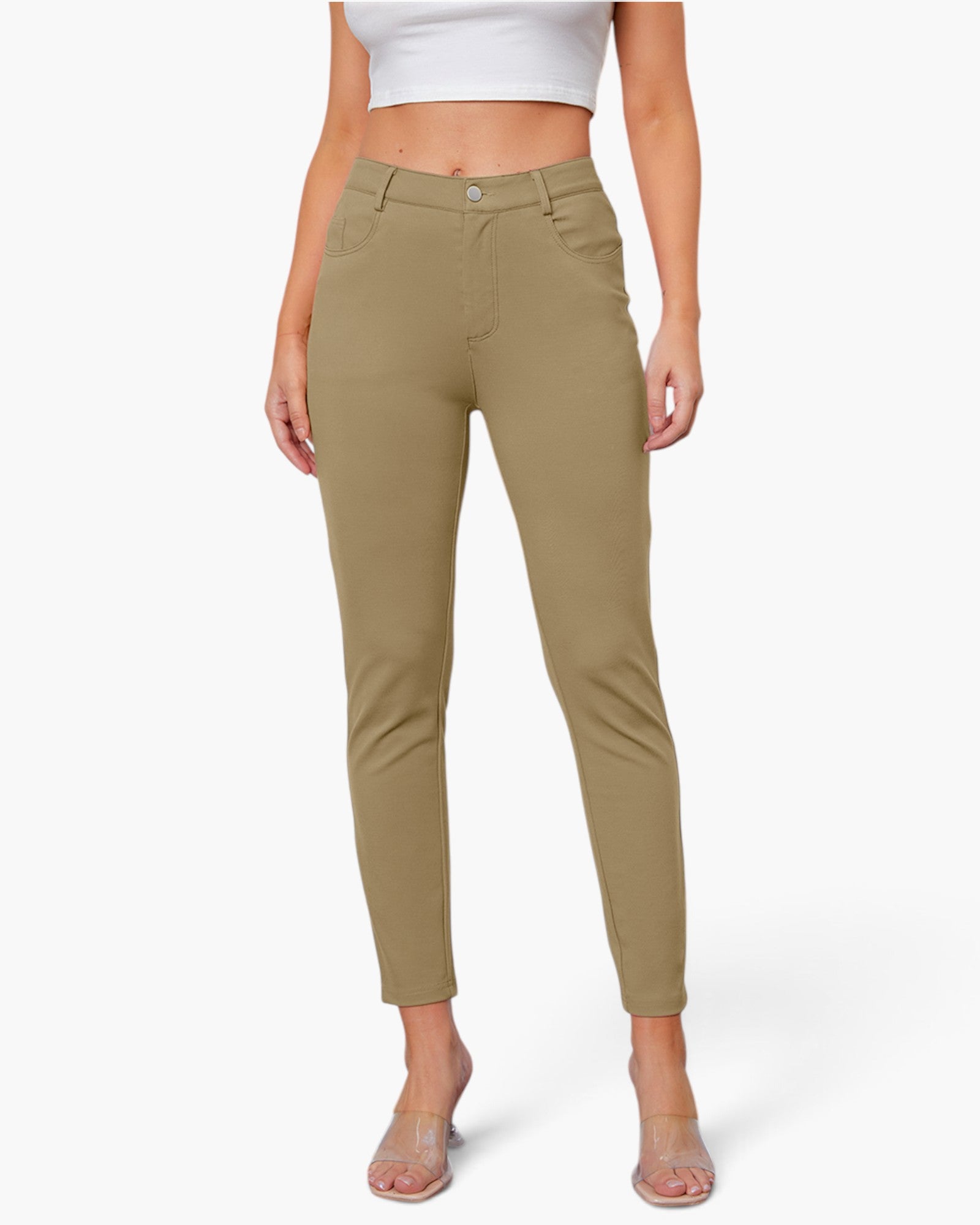 A model wearing slim-fit, ankle-length khaki pants with a white crop top and clear high-heeled sandals, shown against a plain light gray studio background.