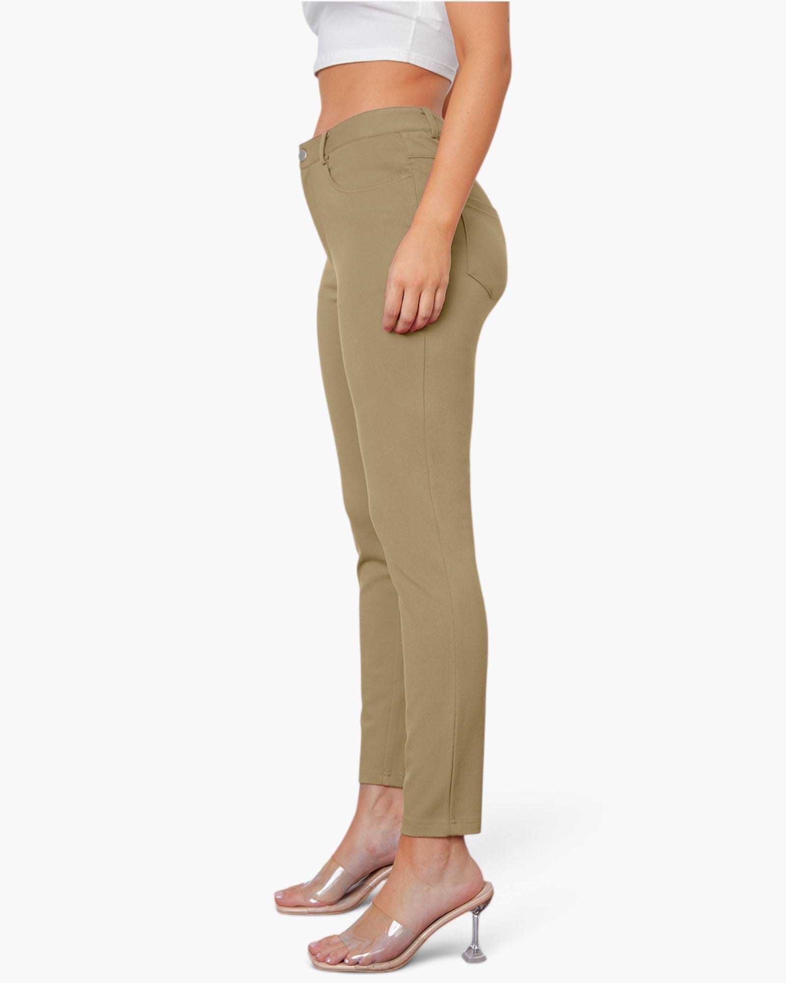 A side-profile view of a model wearing slim-fit, ankle-length khaki pants with a white cropped top and clear high-heeled mule sandals against a plain white background.