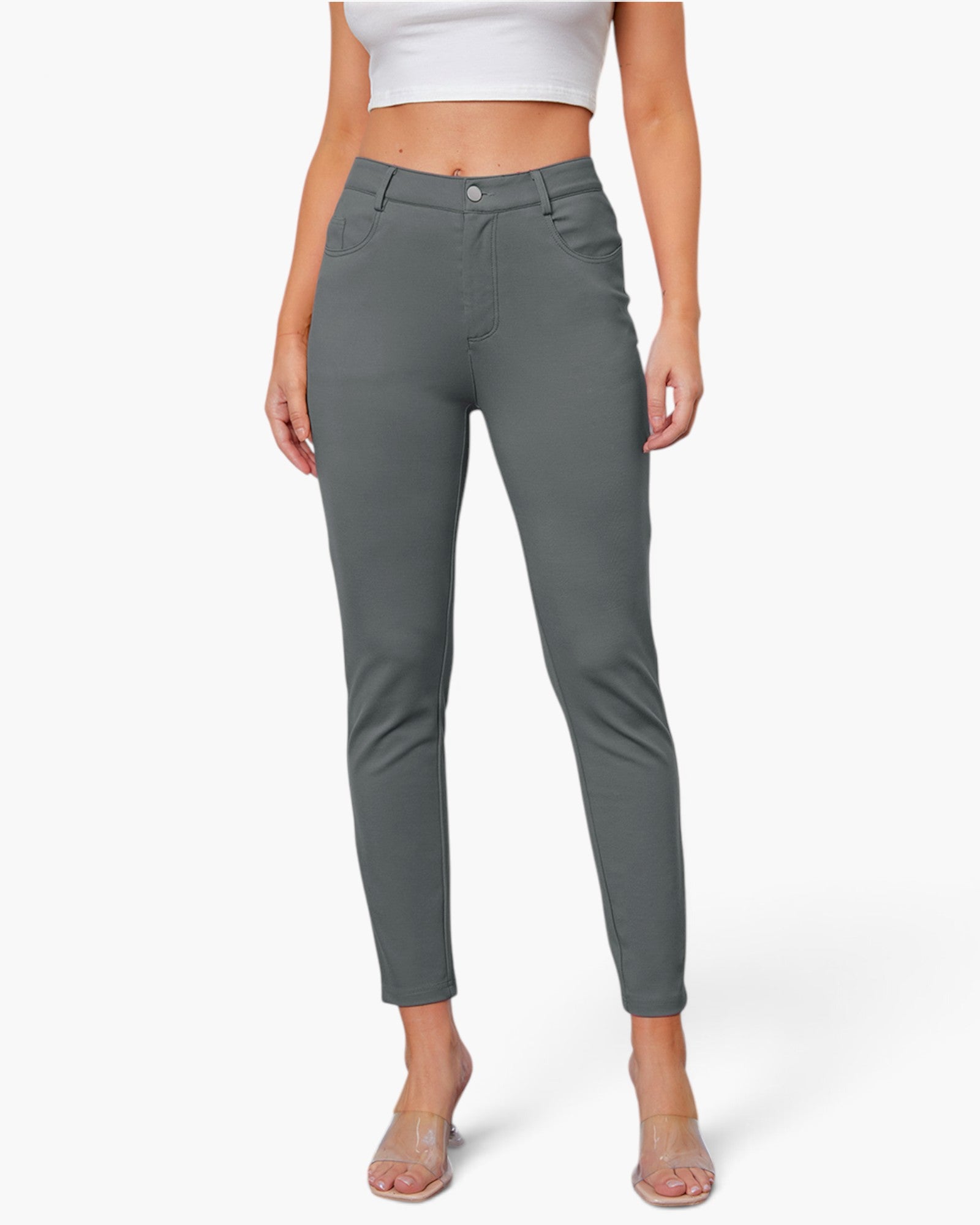 A model wearing a pair of slim-fit, ankle-length gray pants with a button closure and belt loops, paired with a white crop top and clear high-heeled sandals, set against a plain light gray background.
