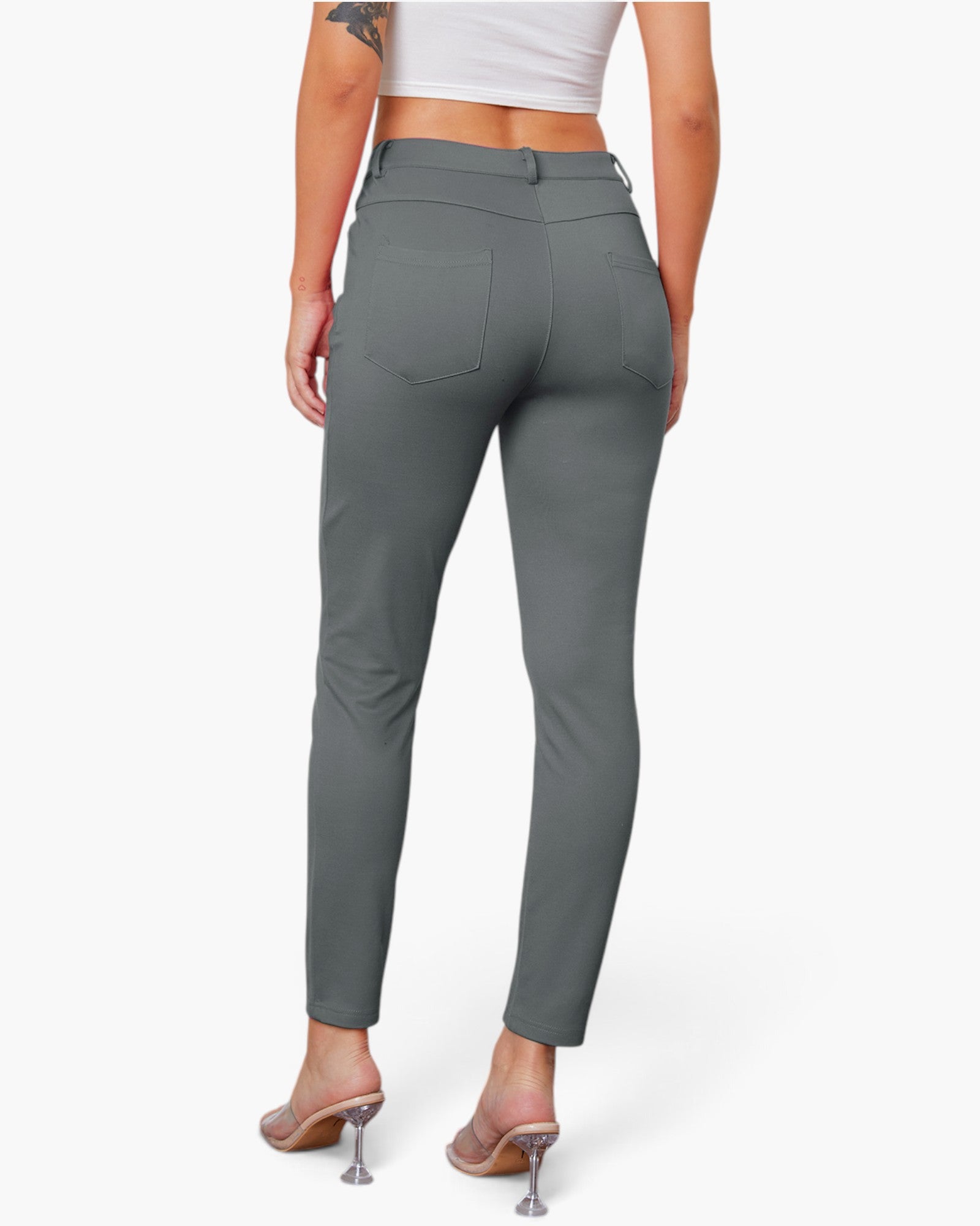 A model shown from the back wearing slim-fit gray ankle-length pants with a belt loop, rear patch pockets, and a button closure, paired with clear high-heel mules against a light gray studio background.