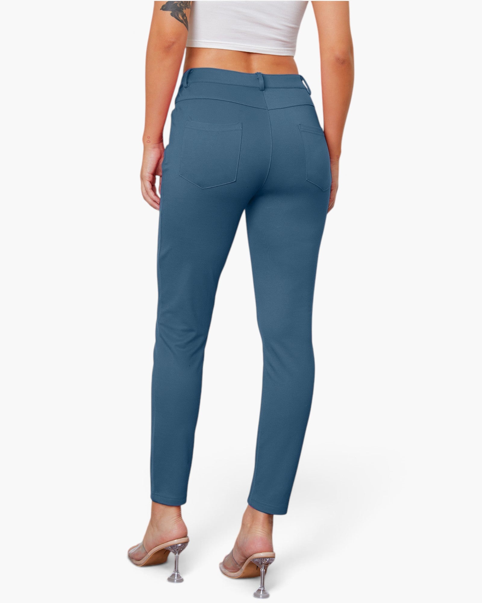 A model shown from the back wearing slim-fit, high-waisted blue pants with a button closure and back pockets, paired with a white crop top and clear high-heel mules, against a light gray studio background.