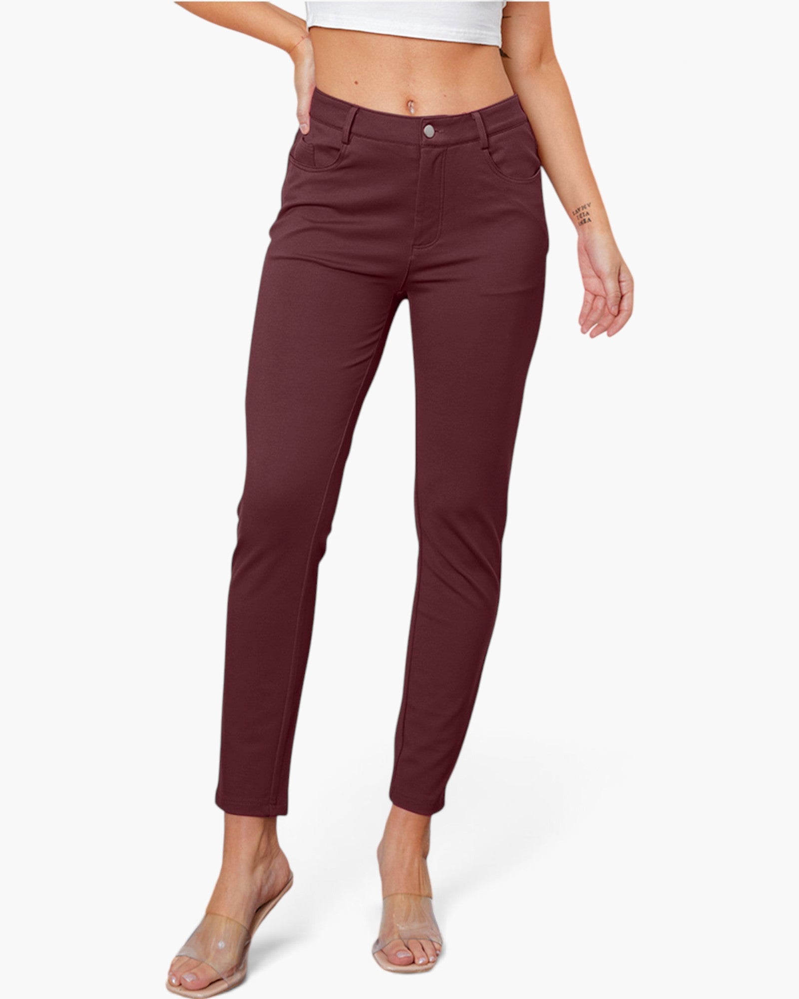 A model wearing slim-fit brown ankle-length pants, paired with a white crop top and clear heeled sandals, against a light gray studio background.