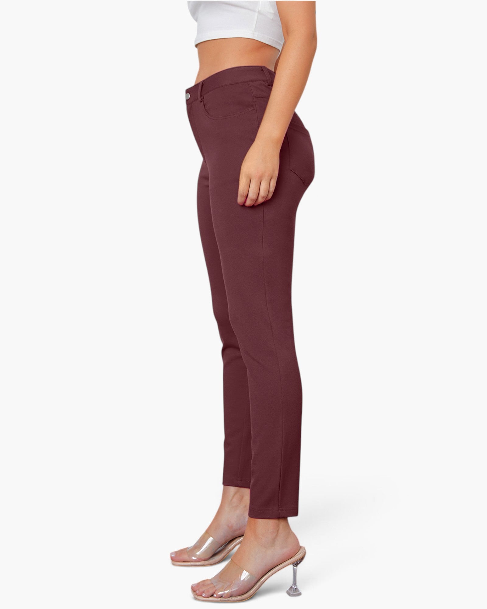 A side-profile view of a model wearing high-waisted, ankle-length slim-fit pants in a deep burgundy or maroon shade, paired with clear high-heel mules and a white crop top, against a plain white studio background.