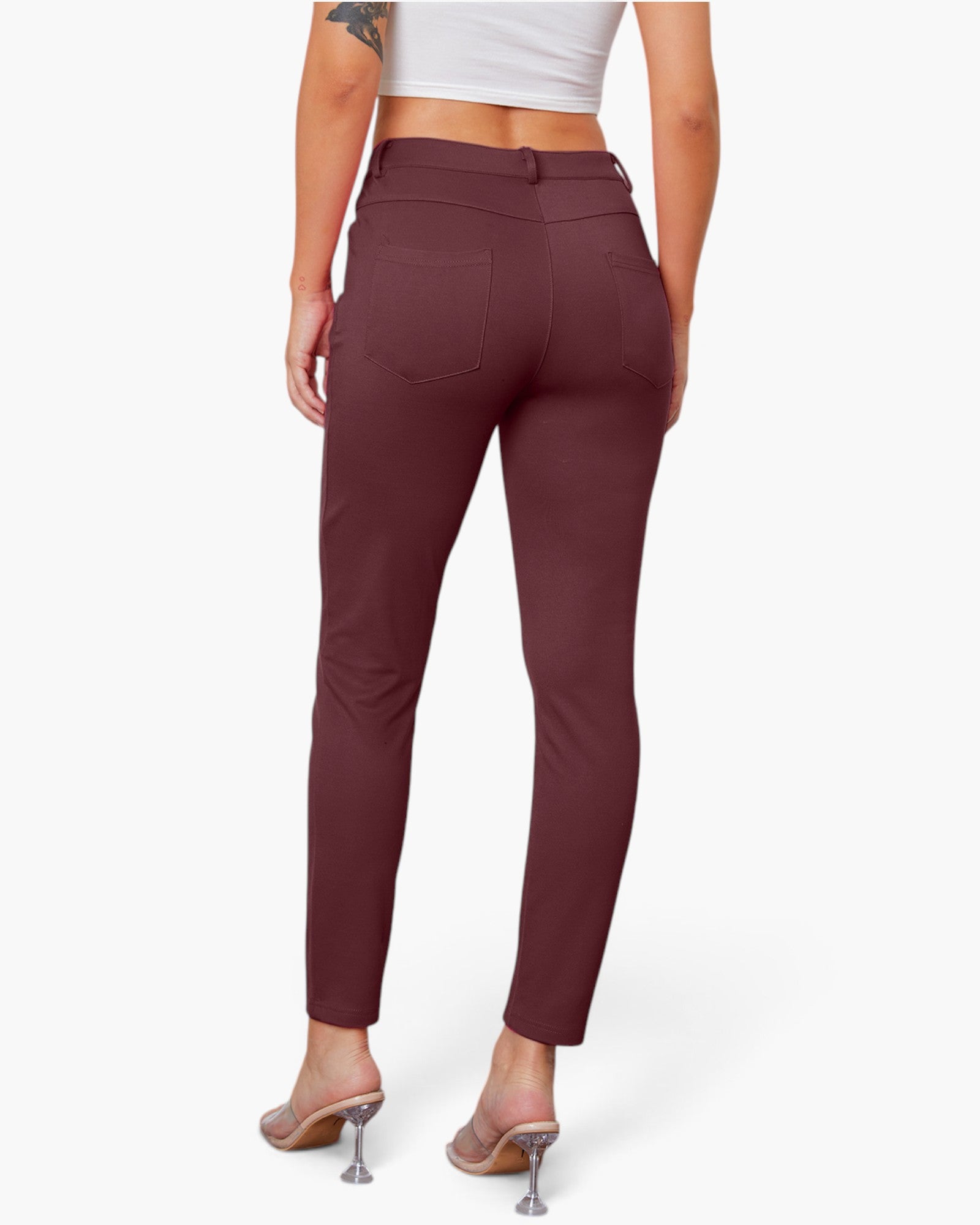 Rear view of a model wearing high-waisted, slim-fit burgundy pants with a button closure and back pockets, paired with a white crop top and clear high-heel mules, against a light gray studio background.