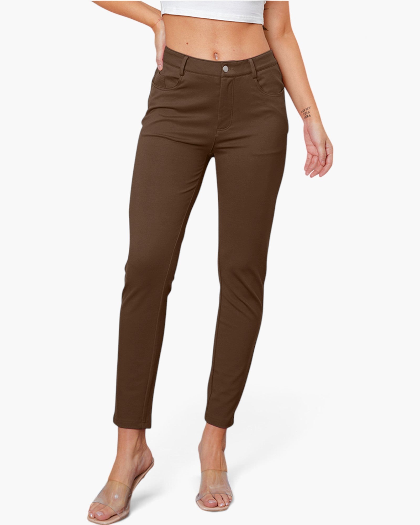 A model wearing slim-fit brown ankle-length pants, paired with a white crop top and clear heeled sandals, against a light gray studio background.