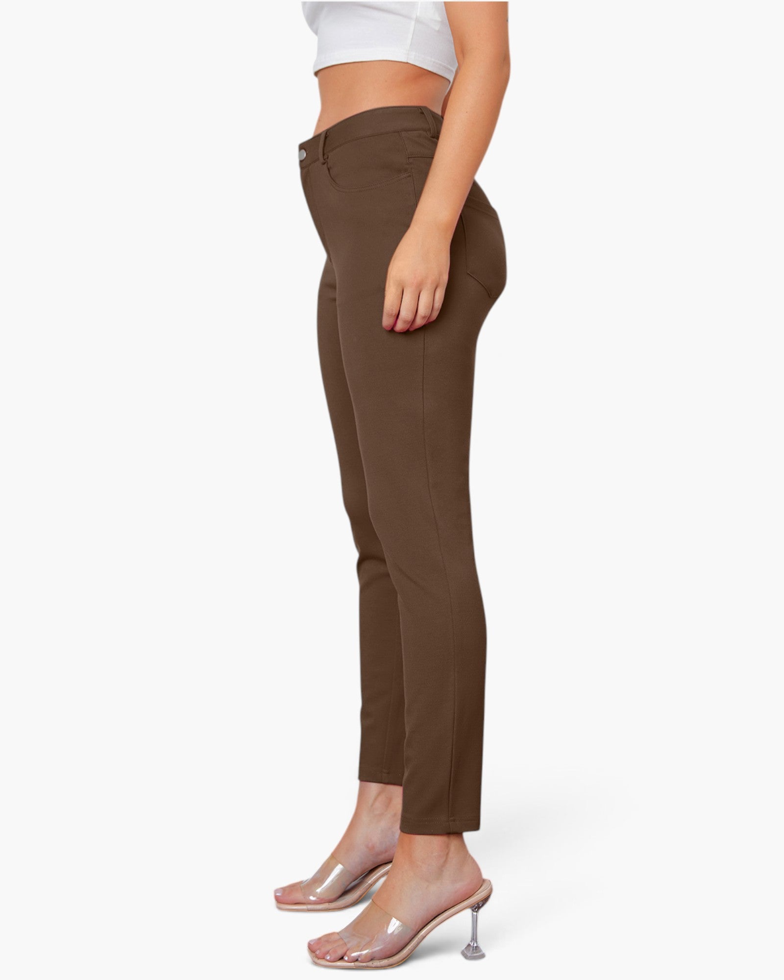 A side-profile view of a model wearing slim-fit brown ankle-length pants, paired with a white crop top and clear high-heeled mule sandals, against a plain white background.