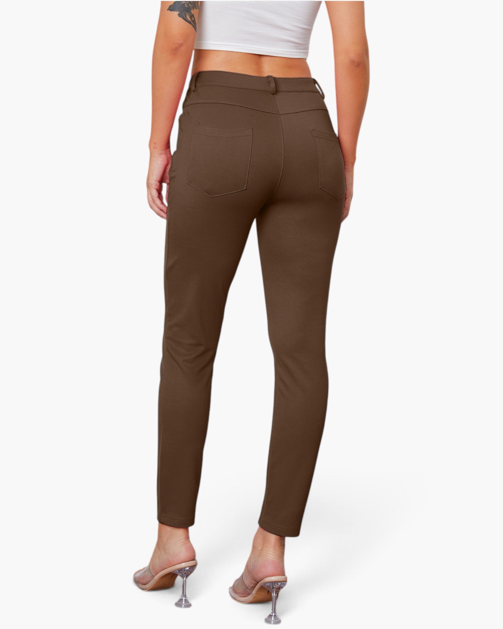 A model shown from the back wearing slim-fit brown ankle-length pants, a white crop top, and clear high-heel mules against a light gray studio background.