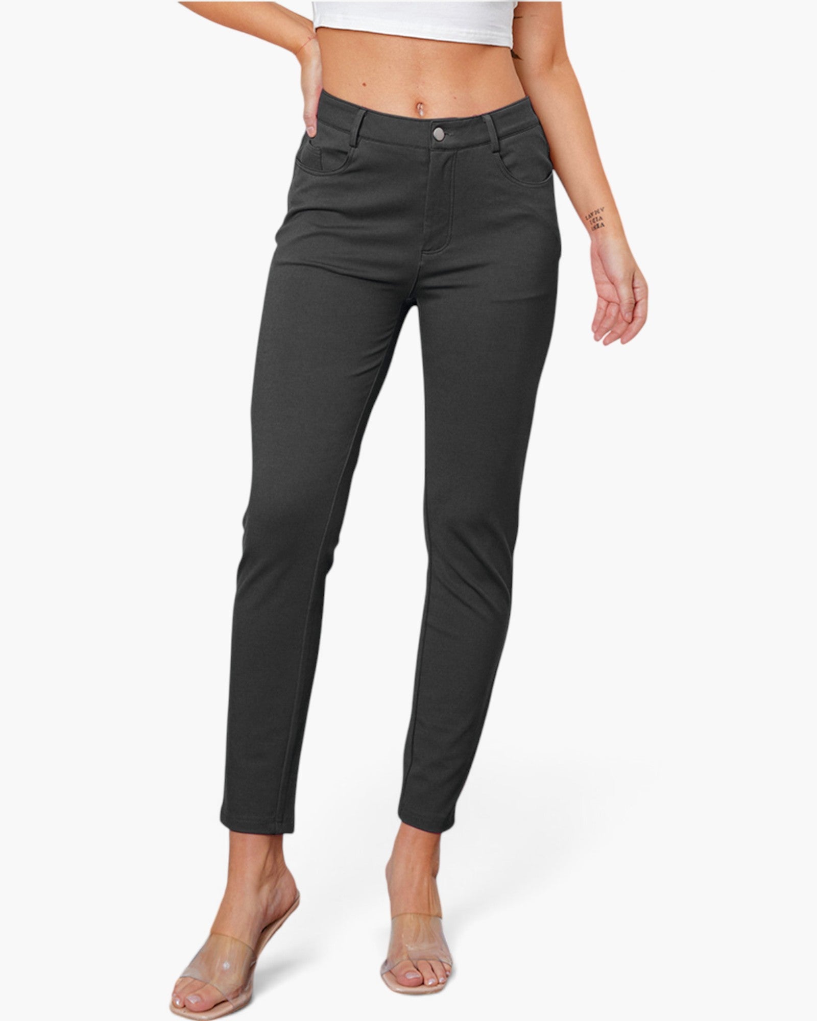A model wearing a pair of dark charcoal gray slim-fit ankle-length jeans, styled with a white crop top and clear heeled sandals against a light gray studio background.