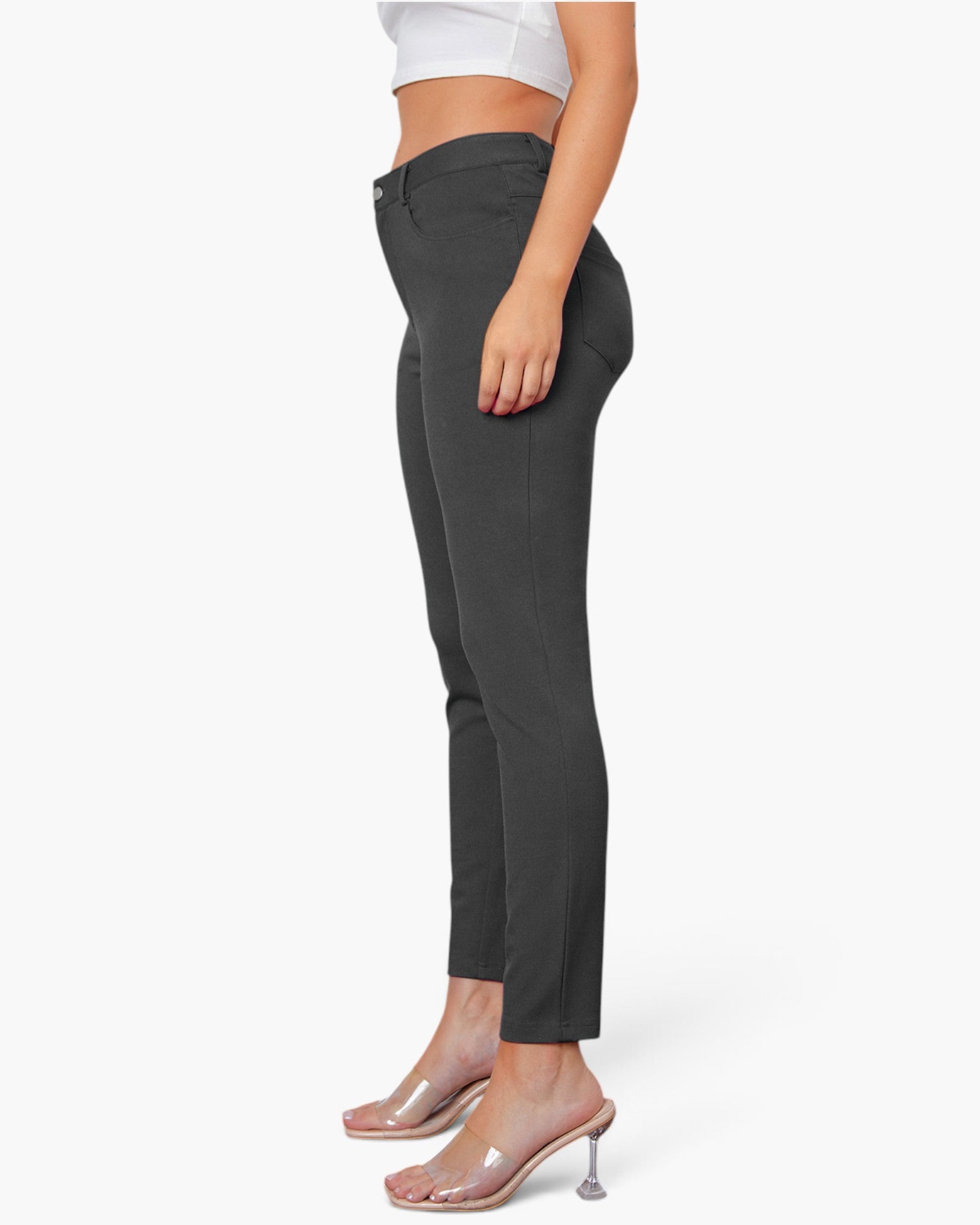 A side-profile view of a model wearing dark charcoal gray slim-fit ankle-length pants, paired with a white crop top and clear high-heeled mule sandals, against a plain white studio background.