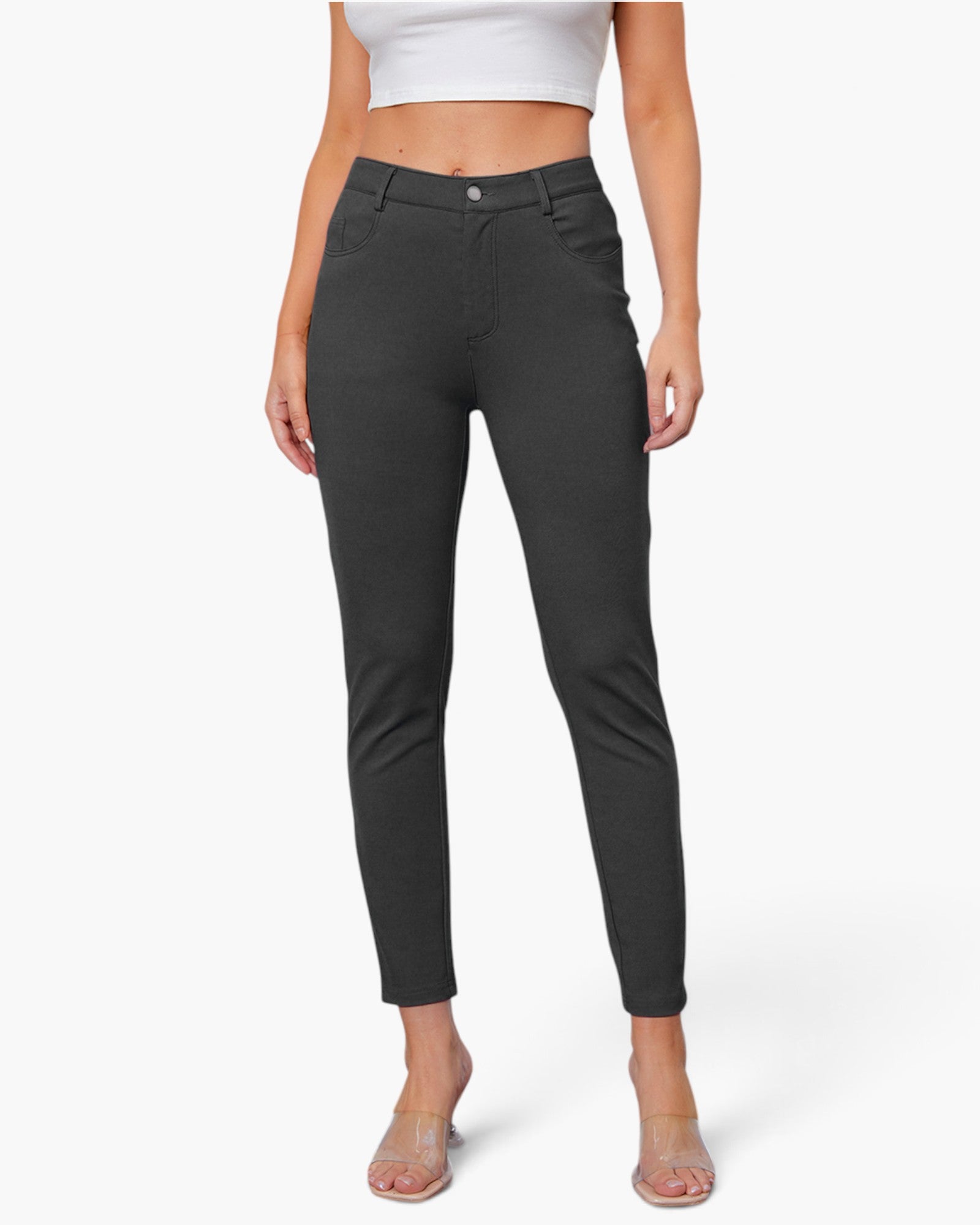 A model wearing a pair of dark charcoal gray slim-fit ankle-length pants, paired with a white crop top and clear high-heeled sandals, against a light gray studio background.