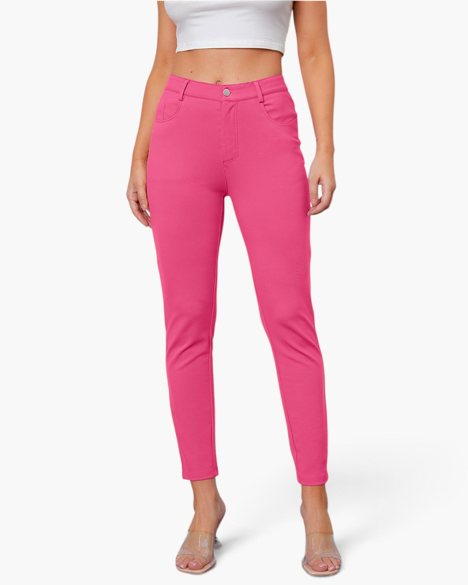 A model wearing bright pink slim-fit ankle-length pants with a white crop top and clear high-heeled sandals, shown against a light gray studio background.