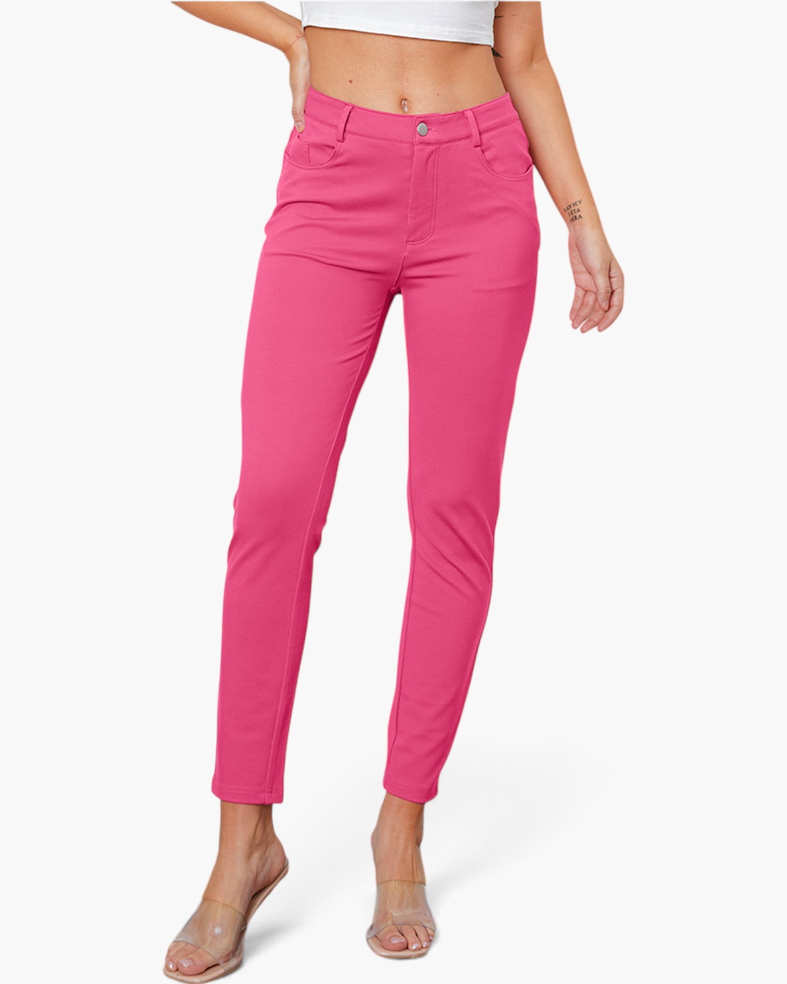 A model wearing bright pink slim-fit ankle-length pants, paired with a white crop top and clear heeled sandals, against a light gray studio background.