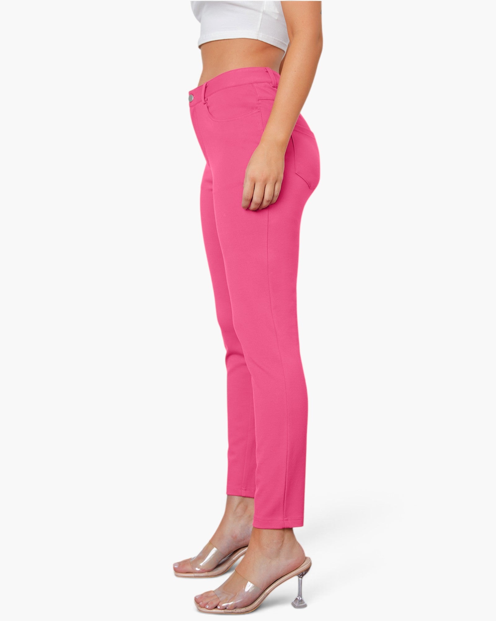 A model shown from the waist down wearing bright pink slim-fit ankle-length pants, a white crop top, and clear high-heeled mule sandals against a plain white background.