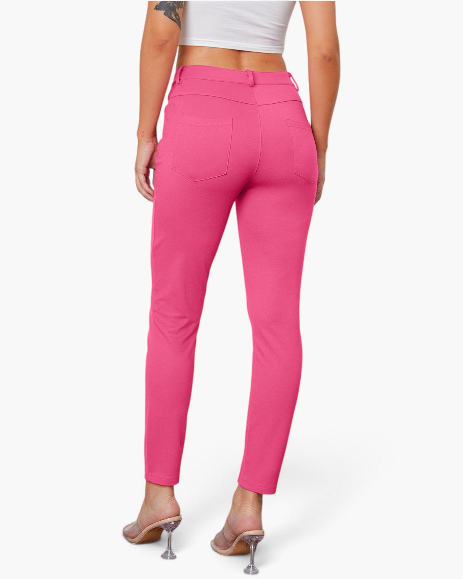 A model shown from the back wearing bright pink slim-fit ankle-length pants with a button waistband and back pockets, paired with a white crop top and clear high-heel mules, against a light gray studio background.