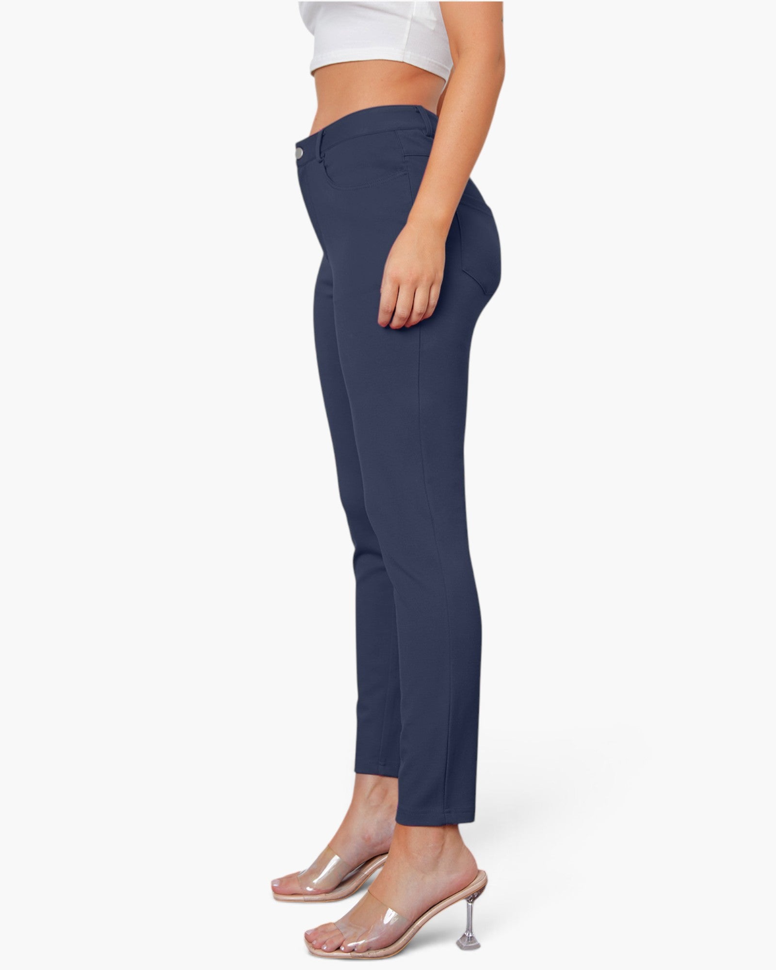 A side-profile view of a model wearing navy blue ankle-length pants, a white crop top, and clear high-heeled mule sandals against a plain white studio background.