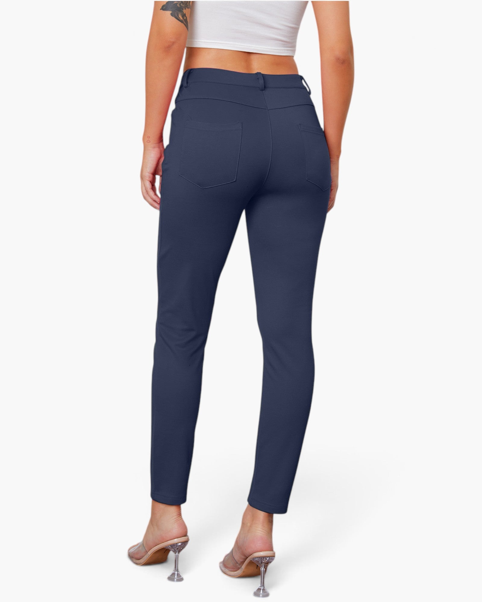 A model shown from the back wearing navy blue slim-fit ankle-length pants, a white crop top, and clear high-heel mules against a light gray studio background.