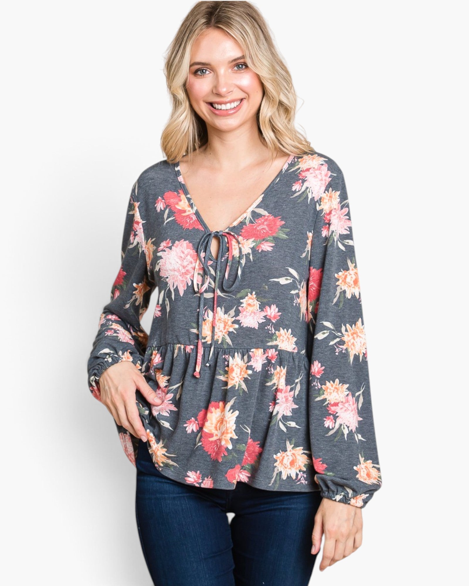 A woman wearing a long-sleeve floral tie-front top in dark gray with pink, red, and peach flowers, paired with dark blue jeans, against a plain white background.