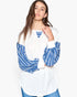 A woman modeling a white long-sleeve top with blue and white striped balloon sleeves and a blue striped V-neckline detail, paired with blue jeans against a plain white background.