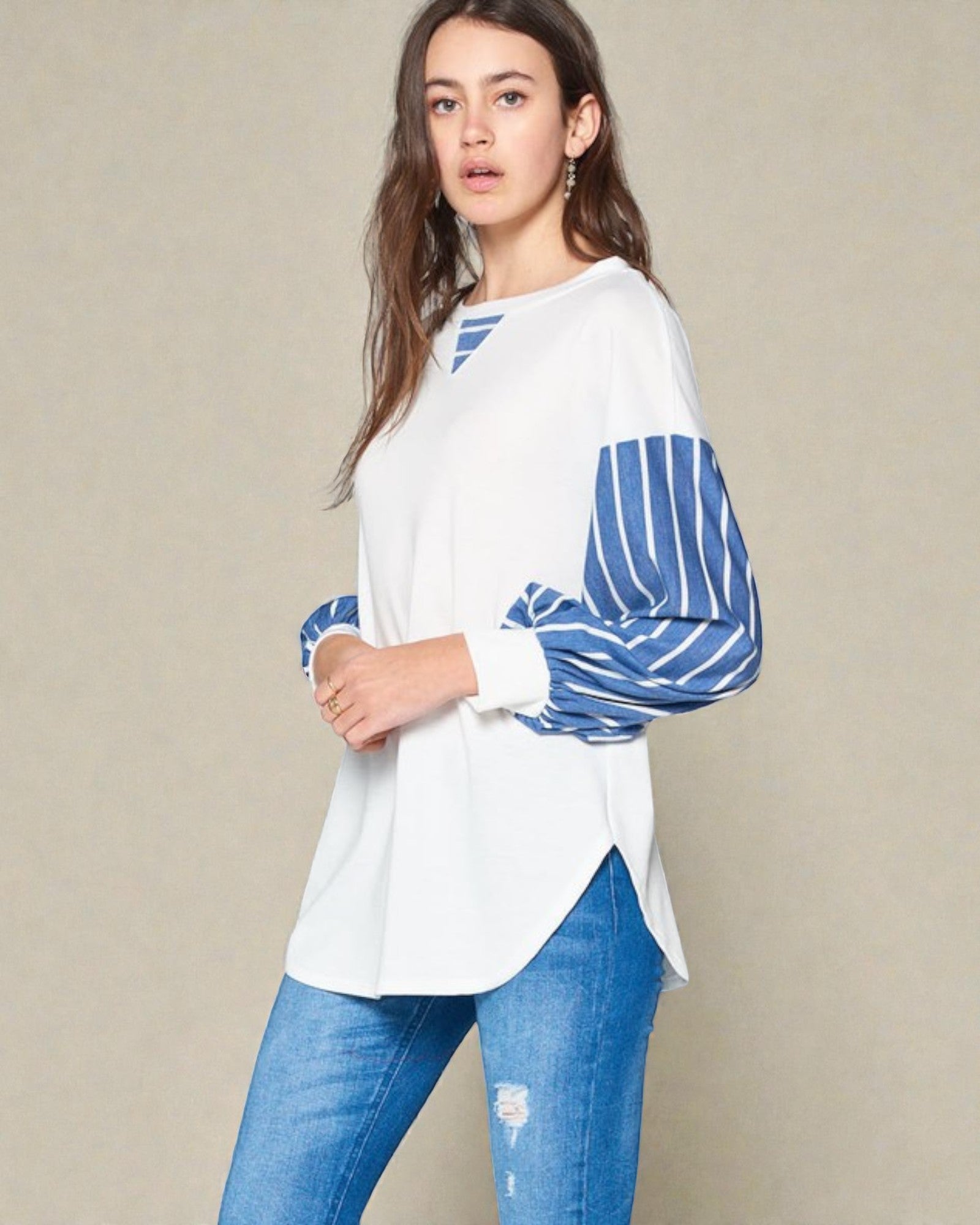 Woman wearing a white top with blue striped sleeves and blue jeans on a beige background