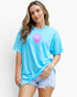 A woman modeling a bright light blue t-shirt with a pink seashell graphic on the chest, paired with light-wash denim shorts, against a plain white background.